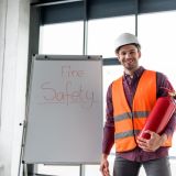 happy-fireman-holding-red-extinguisher-while-standing-near-white-board-with-fire-safety-lettering-e1706457123134.jpg